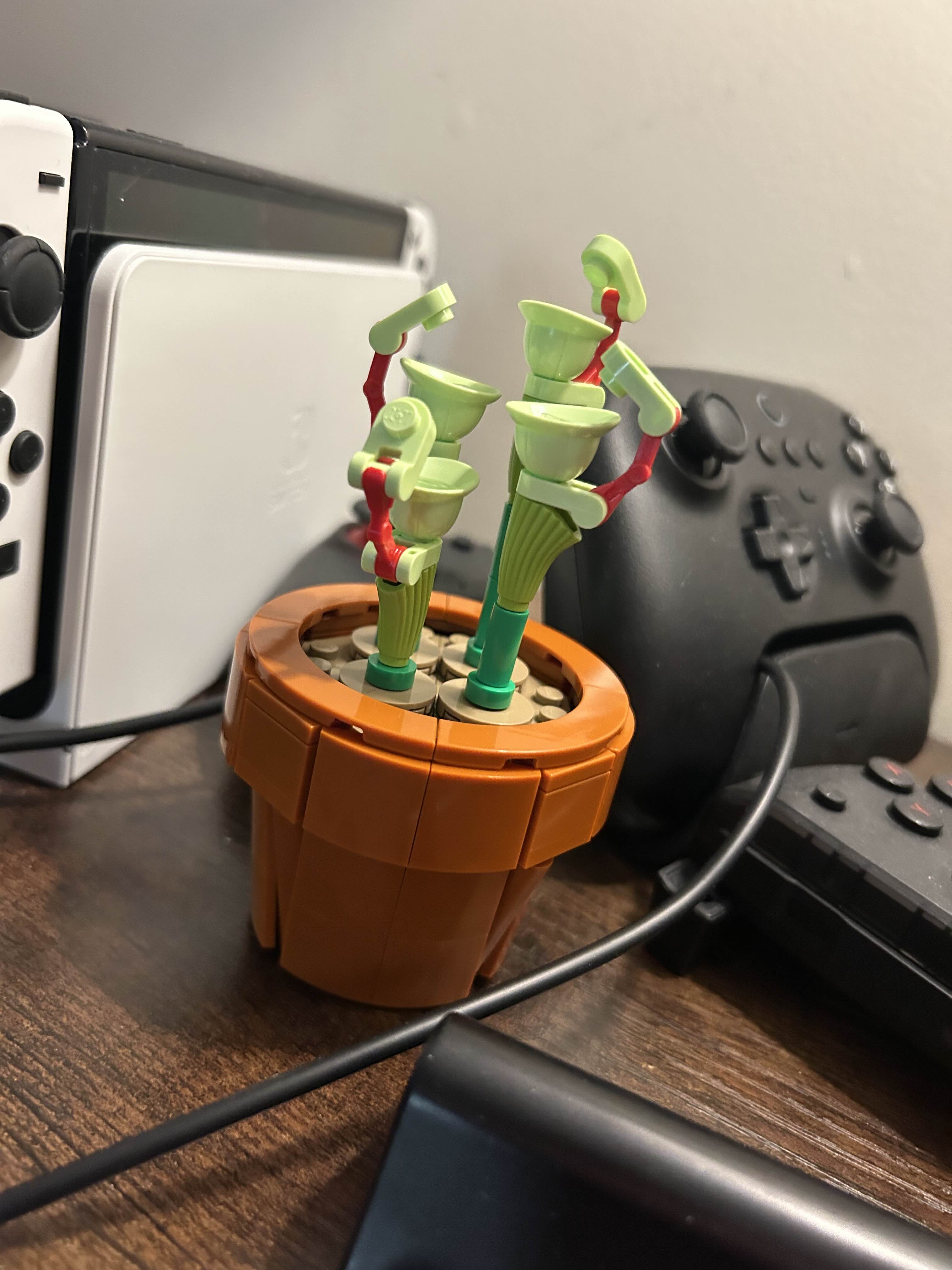 Image of a LEGO flower sitting on a shelf among various game controllers. It is in a brown pot and is a light green, tall flower with small coverings on the top.